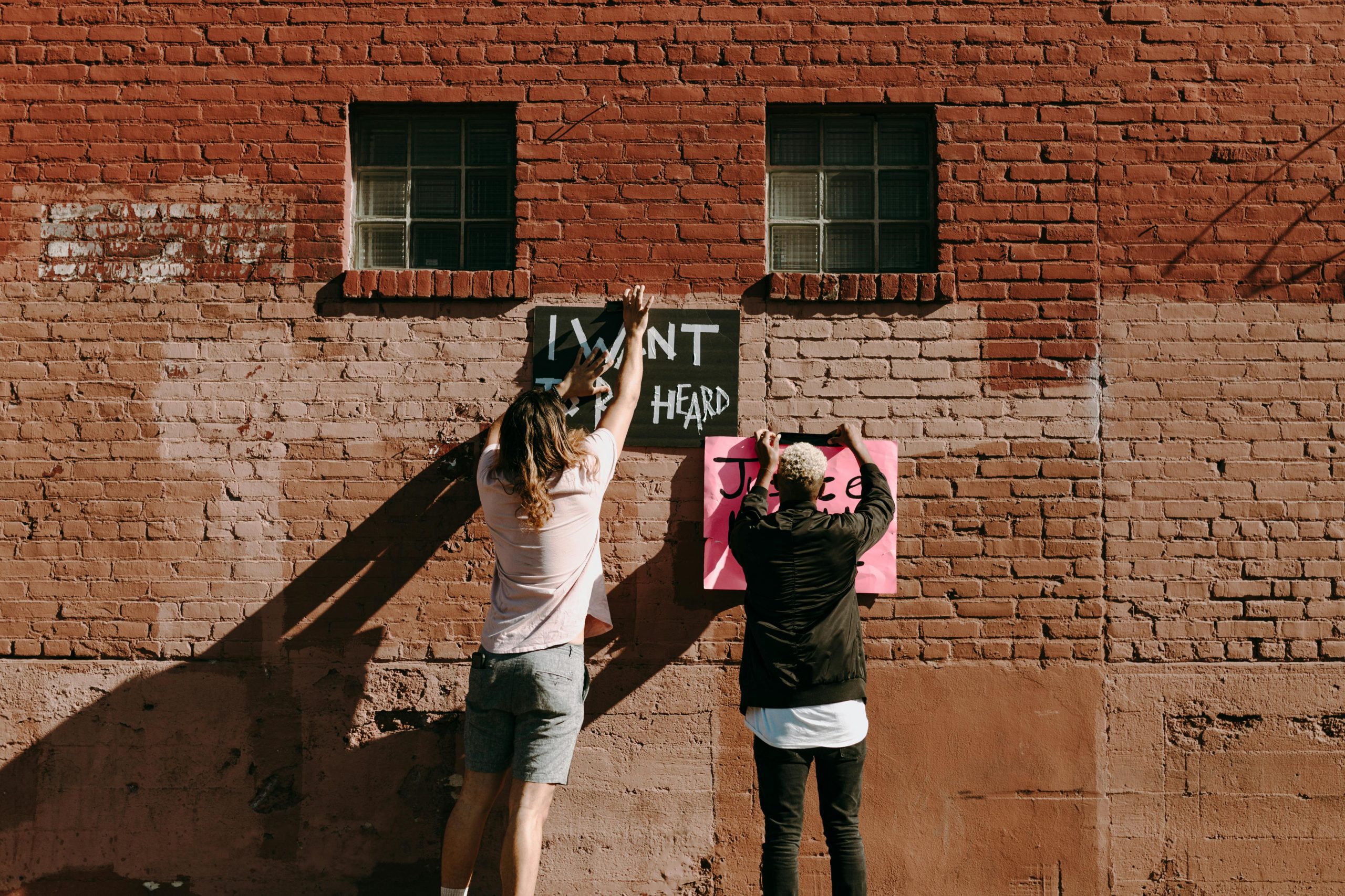 Two individuals placing protest posters on a brick wall, emphasizing justice and being heard.